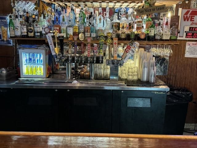 The back bar: shelves of spirits, a bank of draught taps, stacked plastic cups, and a hand-lettered sign reading “Play Nice or Leave”.