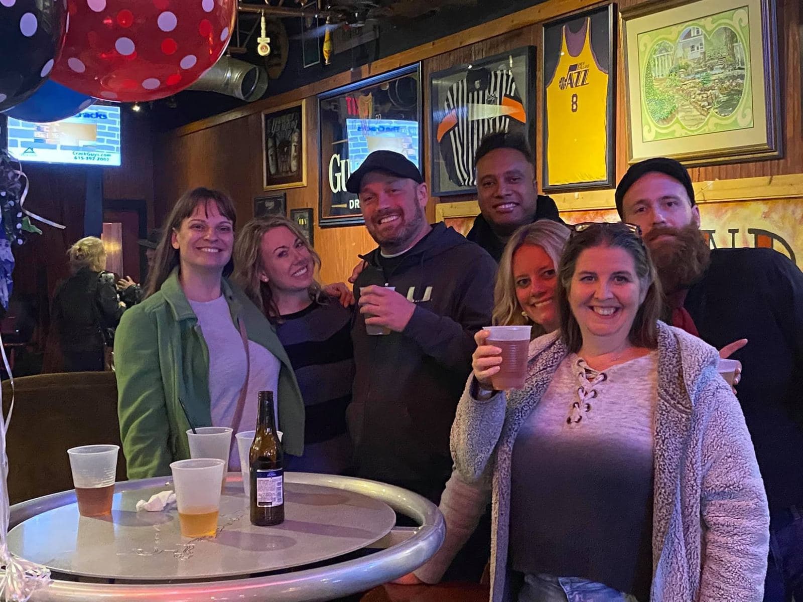 Seven friends crowded around a high-top table raising cups, balloons overhead and framed jerseys on the wall behind them.