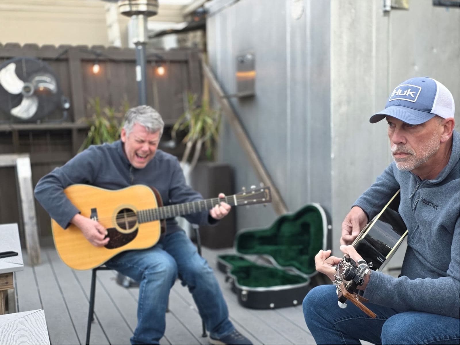 Two guitarists playing on the outdoor patio, one singing, string lights and a patio heater on the fence behind them.