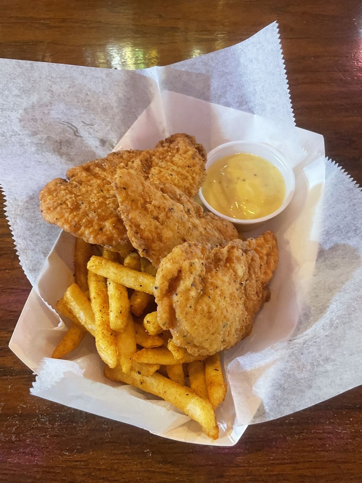 Three breaded chicken tenders and seasoned fries in a paper-lined basket with a cup of dipping sauce.