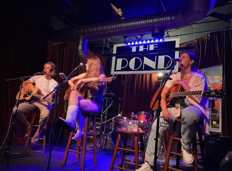 Three musicians on stools with acoustic guitars, playing under blue stage light in front of The Pond’s painted stage sign.